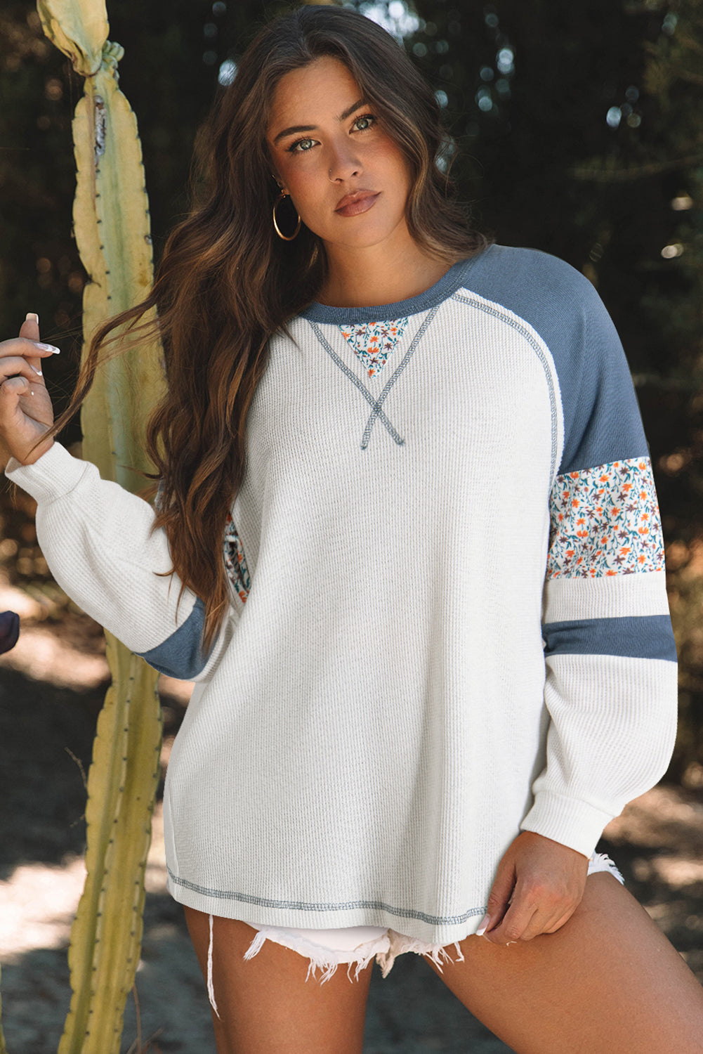 Woman wearing a white and blue sweater with floral sleeves, standing next to a cactus.