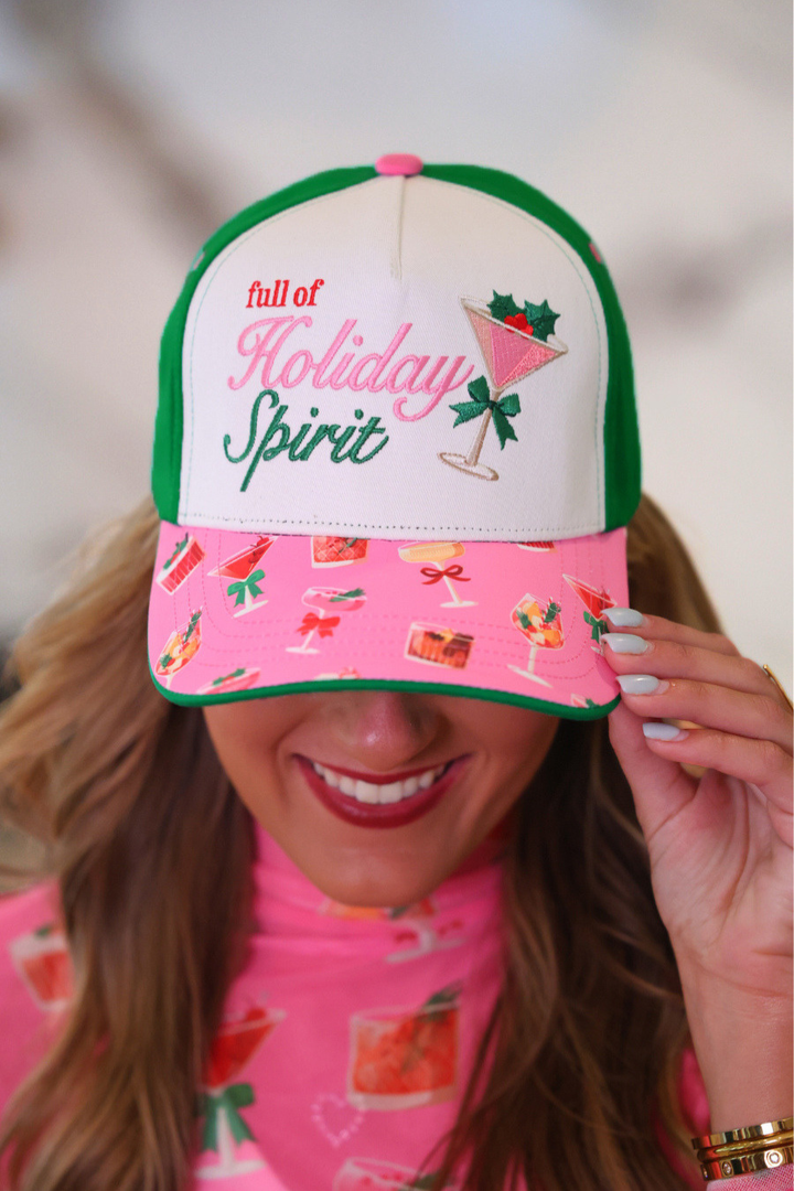A woman wearing a festive Christmas-themed trucker hat with a white crown, green mesh side panels, and a pink brim covered in holiday drink illustrations. The front of the hat features the embroidered text &