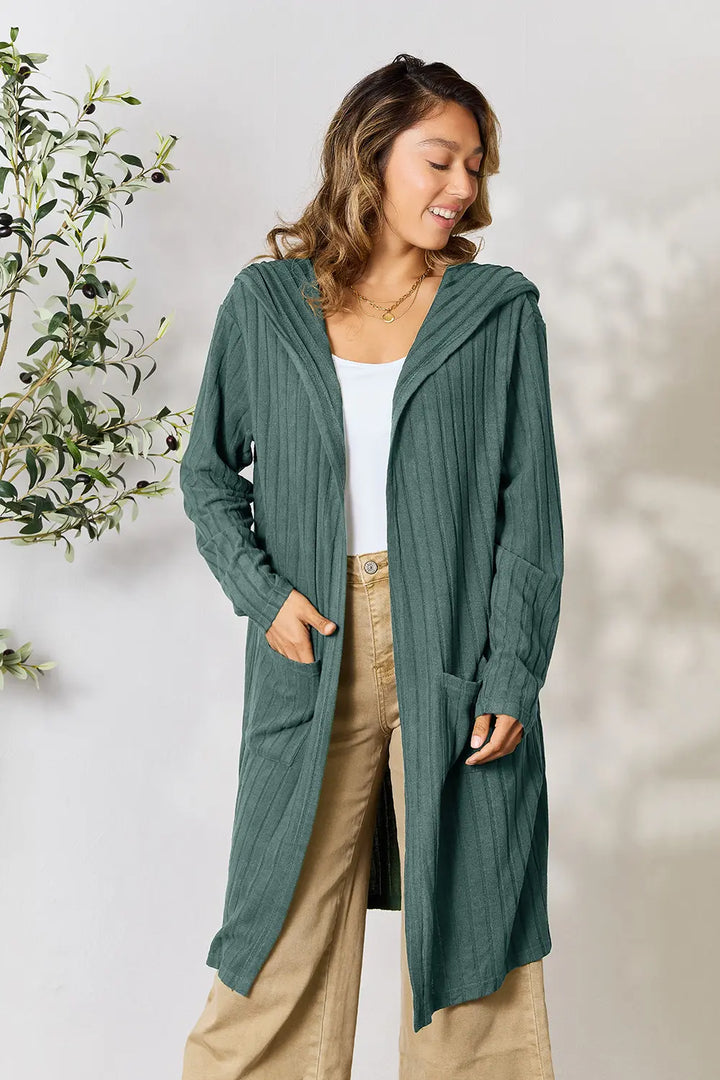 Woman wearing a long green cardigan with a white top and beige pants, standing against a light background.
