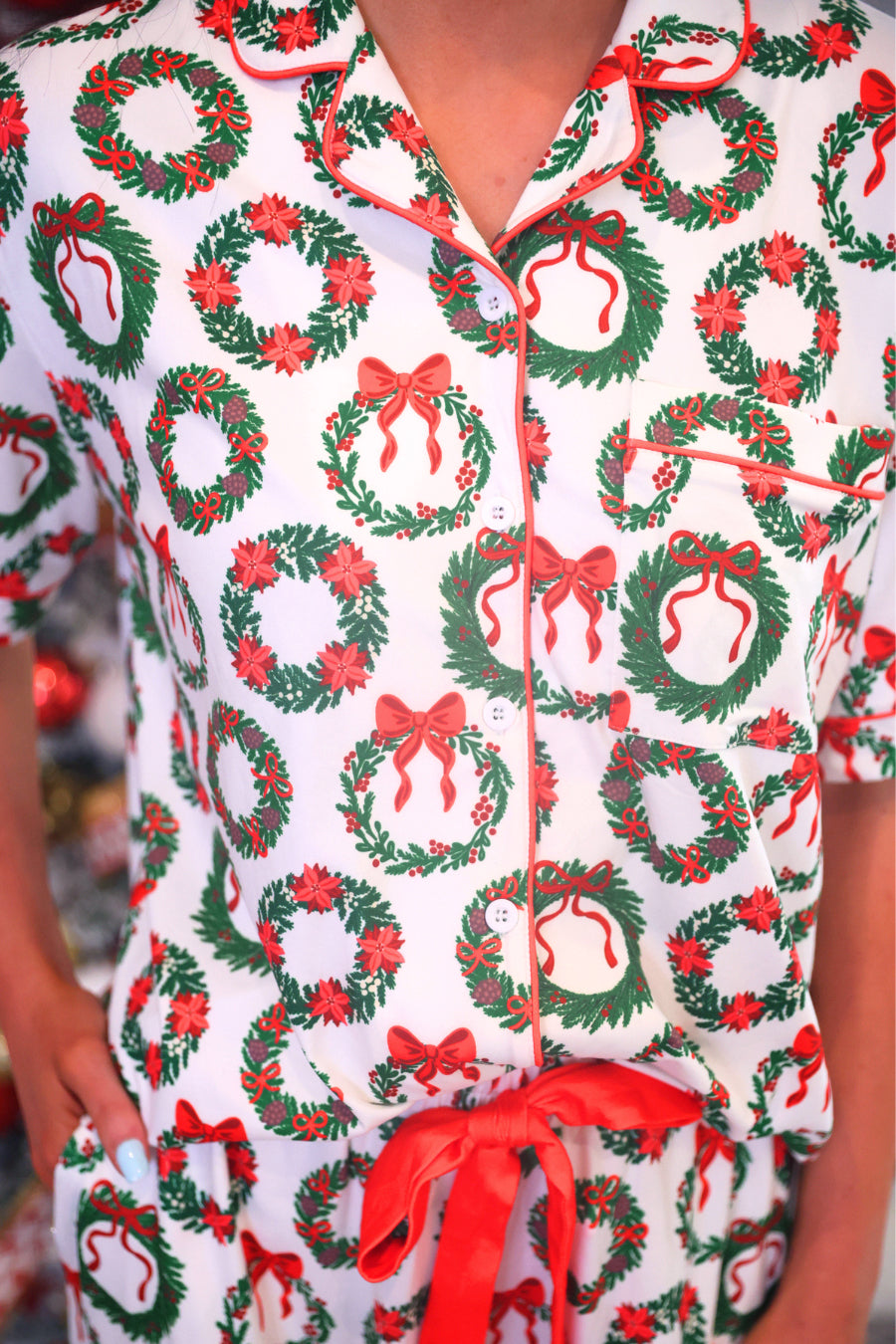 A close-up of a person wearing a white short-sleeve pajama top with a festive Christmas wreath print. The pajamas feature green wreaths with red bows and red trim along the collar, placket, and pocket edges.