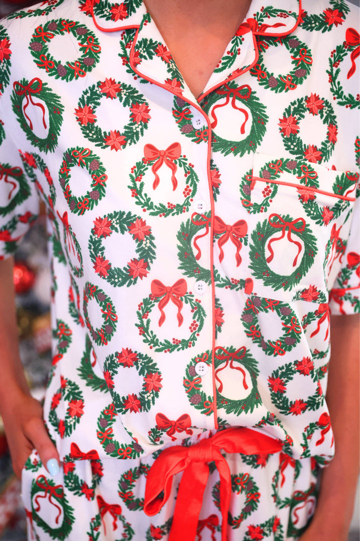 A close-up of a person wearing a white short-sleeve pajama top with a festive Christmas wreath print. The pajamas feature green wreaths with red bows and red trim along the collar, placket, and pocket edges.