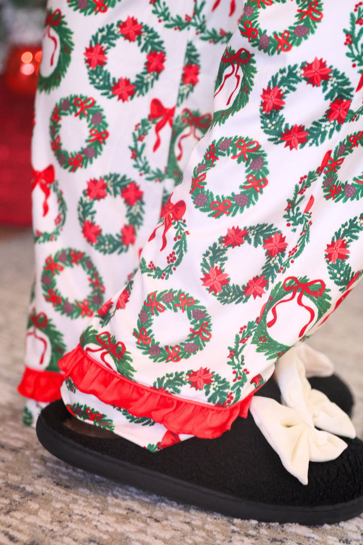 Close-up of a person wearing white holiday pajama pants with a green wreath and red ribbon print, red ruffled hems, and black slip-on slippers with cream bows.