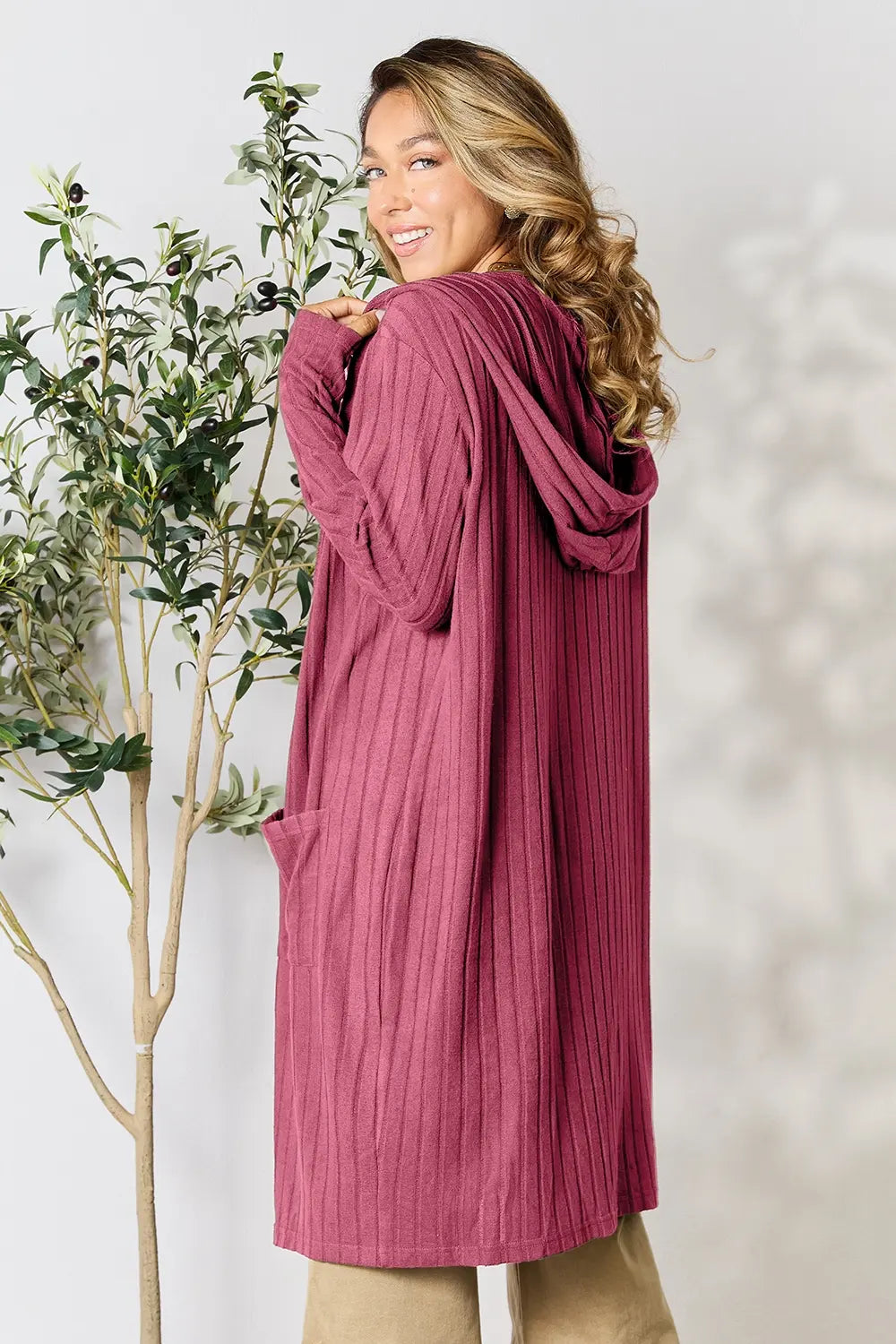 Woman wearing a pink cardigan standing next to a plant against a white background
