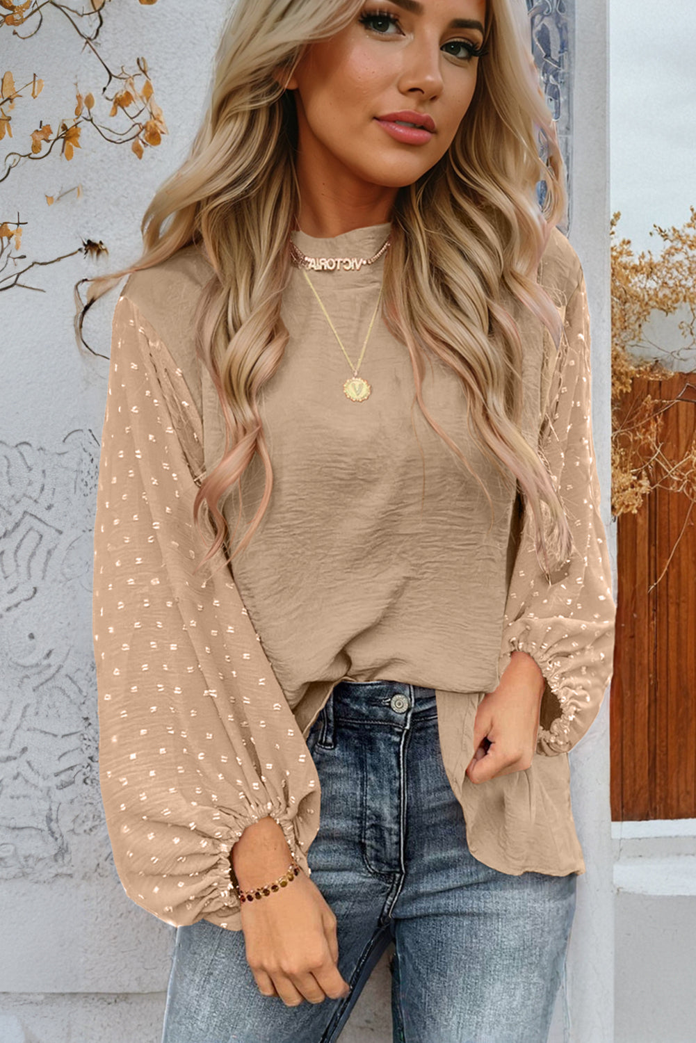 Woman wearing a beige blouse with long sleeves and blue jeans against a decorative wall.