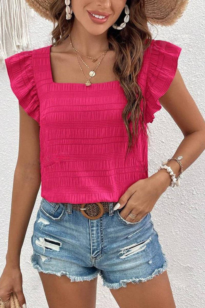 Woman wearing a textured, rose-colored top with ruffled sleeves and square neckline and a pair of jean shorts