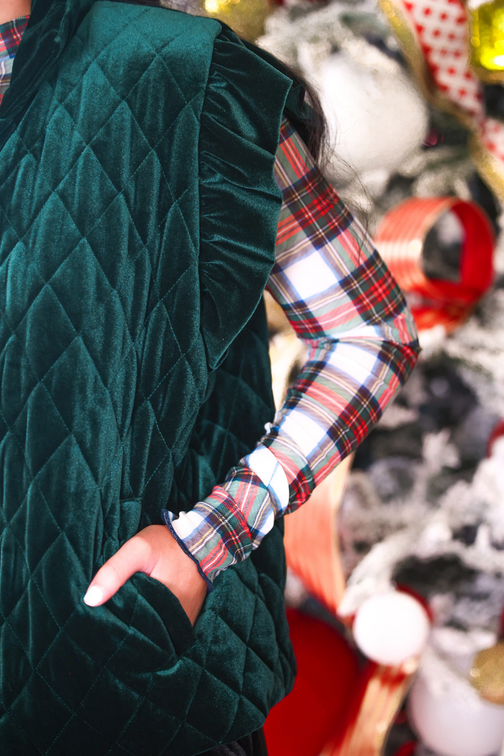 A close-up of a person wearing a dark green quilted velvet vest over a red, green, and white plaid long-sleeve shirt, with a hand in the pocket of the vest, set against a festive blurred holiday background.