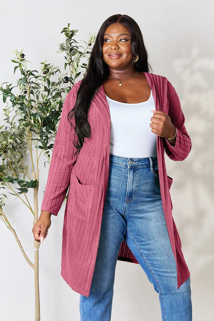 Woman wearing a pink cardigan, white top, and blue jeans standing next to a plant.