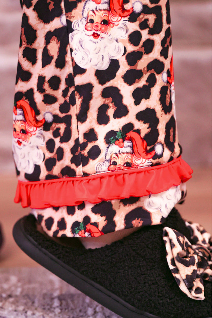 Close-up of a person wearing festive pajama pants with a leopard print pattern, Santa Claus faces, and a red ruffle hem, paired with a black fuzzy slipper featuring a matching leopard print bow.