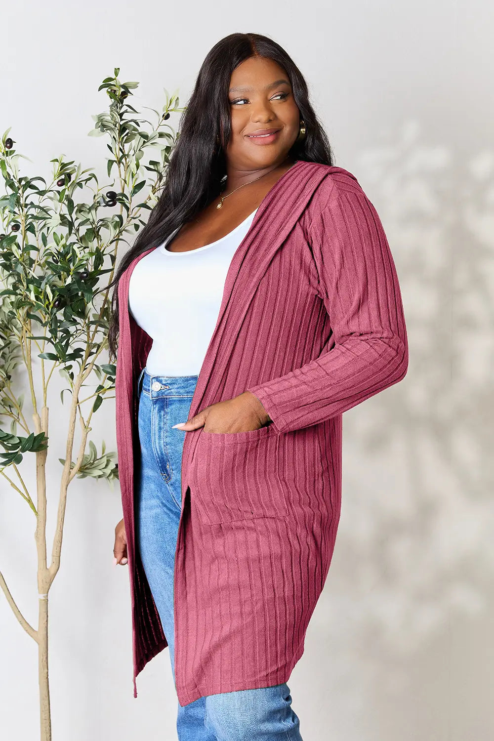 Woman wearing a pink ribbed cardigan over a white top and blue jeans, standing against a neutral background with a plant to the left.