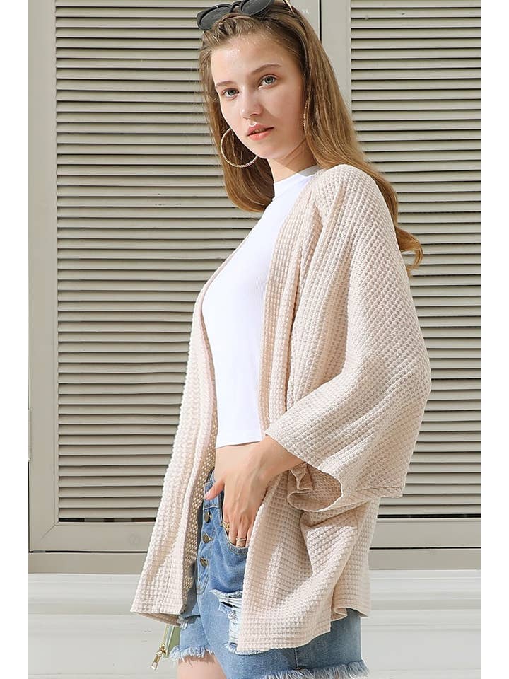Woman wearing a coconut cardigan over a white shirt and denim shorts, standing in front of a neutral background.