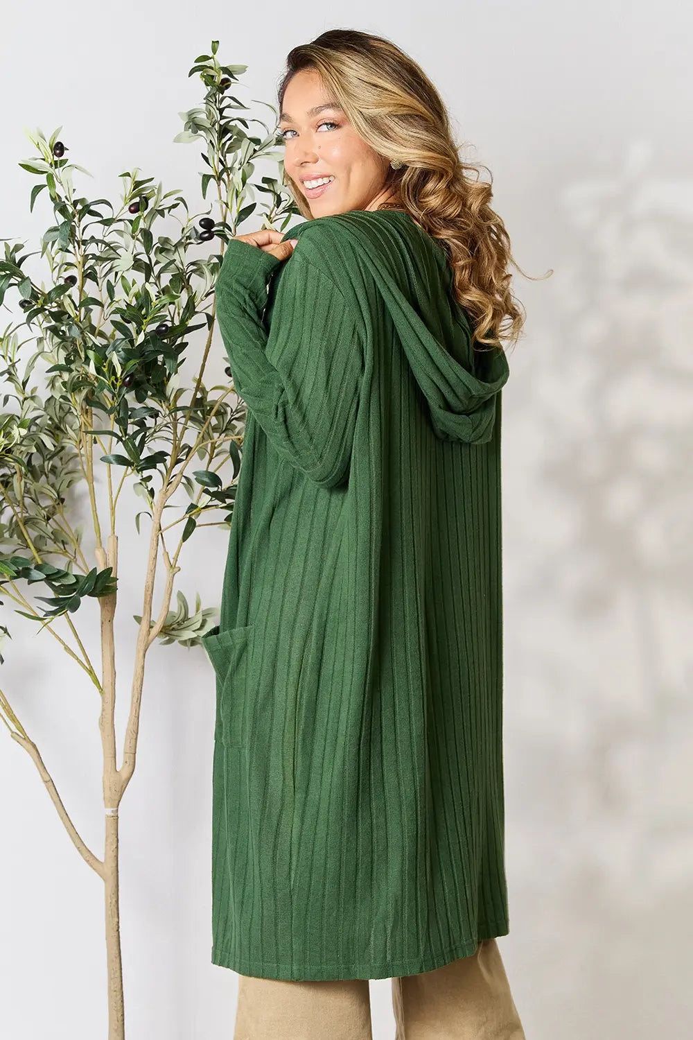 Woman wearing a green cardigan standing next to a plant against a white background