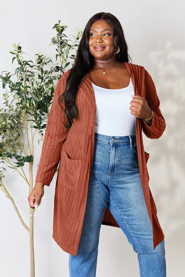 Woman wearing a rust-colored cardigan, white top, and blue jeans standing next to a plant.