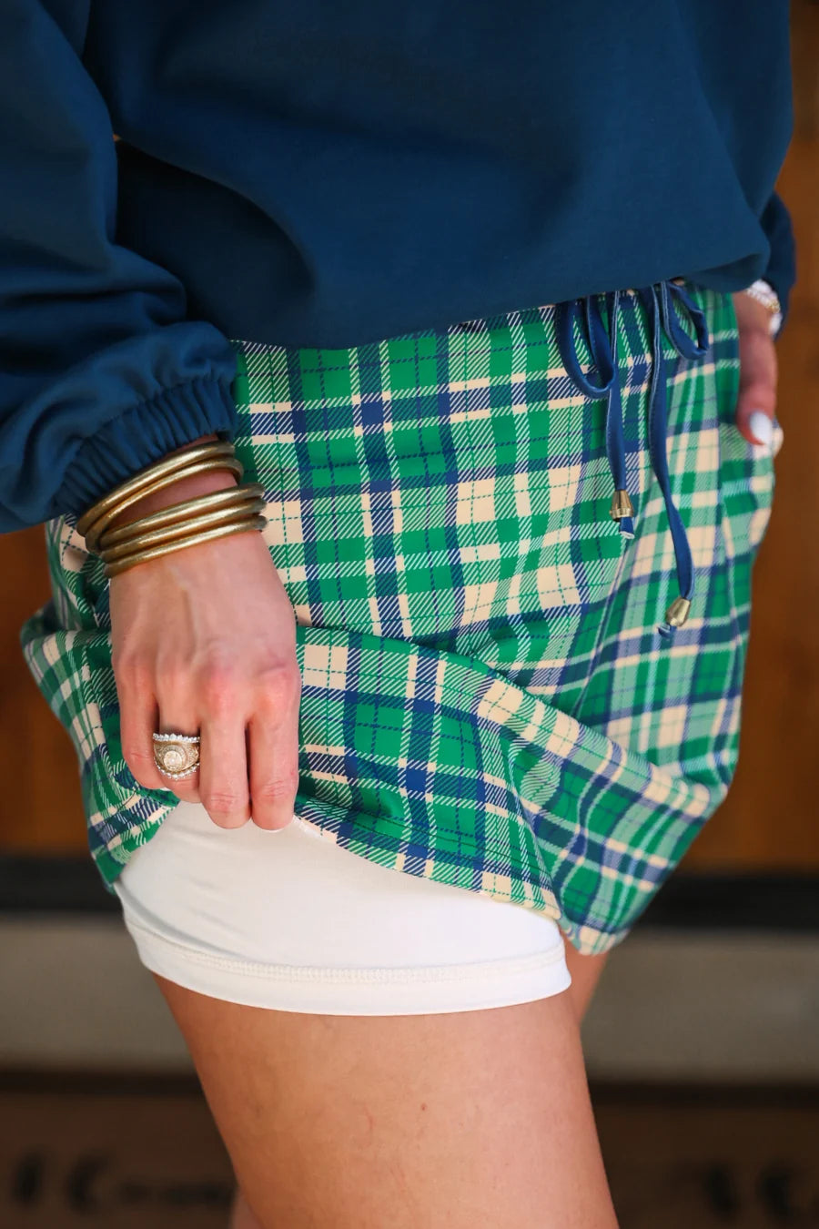 A close-up of a person wearing green, blue, and cream plaid skort with a drawstring, layered over white fitted shorts and a navy blue sweatshirt.