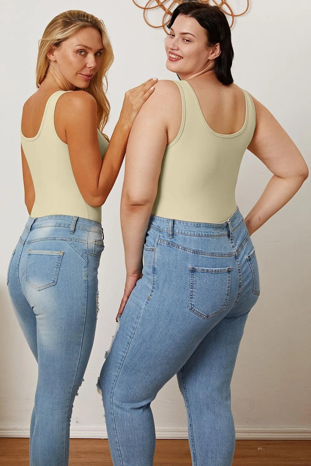 Two women wearing light green tank tops and blue jeans standing against a plain background.