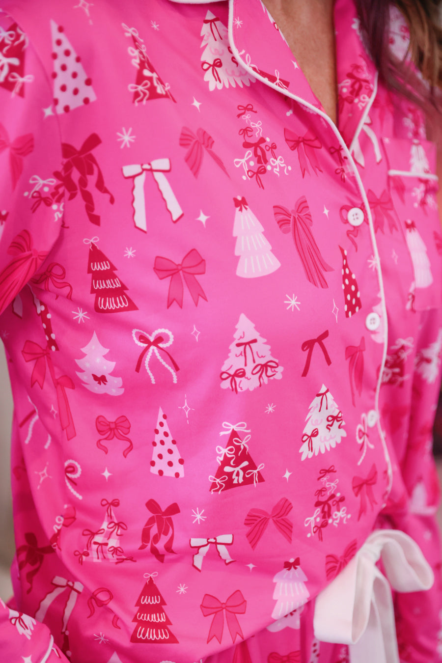 A close-up of a bright pink pajama top with a white and red Christmas print featuring bows, trees, and gifts. A white tie belt is visible at the waist.