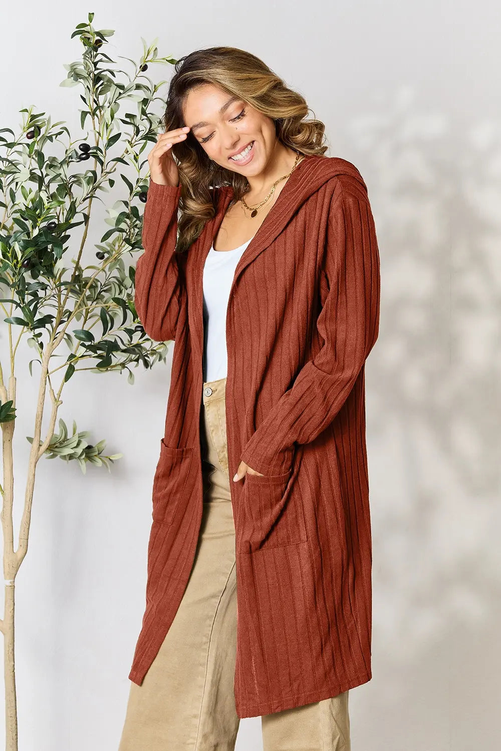 Woman wearing a rust-colored cardigan standing next to a plant against a white wall