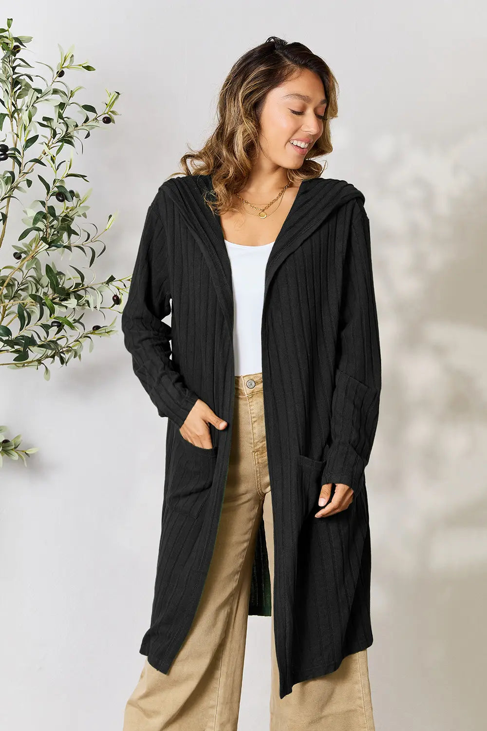Woman wearing a black long cardigan with a white top and beige pants, standing against a light background.