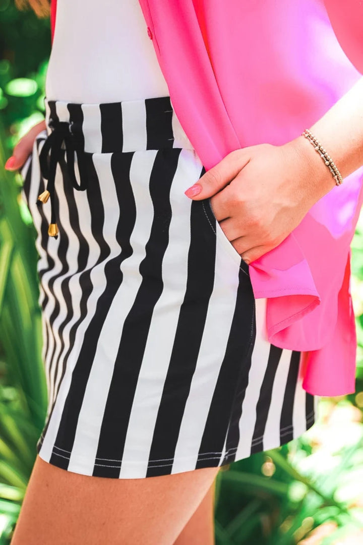 Woman wearing a black and white strips skort with a white tee and pink open blouse, hands in the pockets.