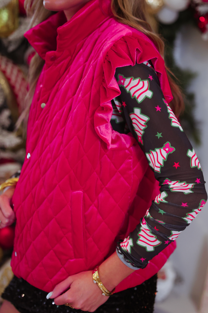 A close-up of a person wearing a bright pink quilted sleeveless vest over a black long-sleeve shirt with a Christmas tree pattern, in a festive indoor setting.