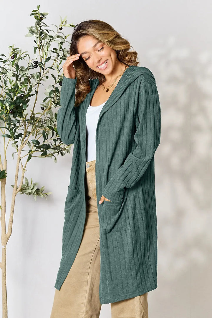 Woman wearing a green cardigan standing next to a plant with a neutral background