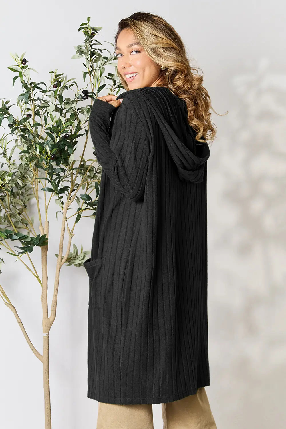 Woman wearing a black cardigan standing next to a plant against a white wall