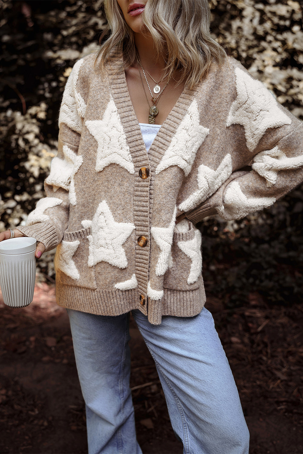 Person wearing a beige cardigan with star patterns, holding a mug outdoors.