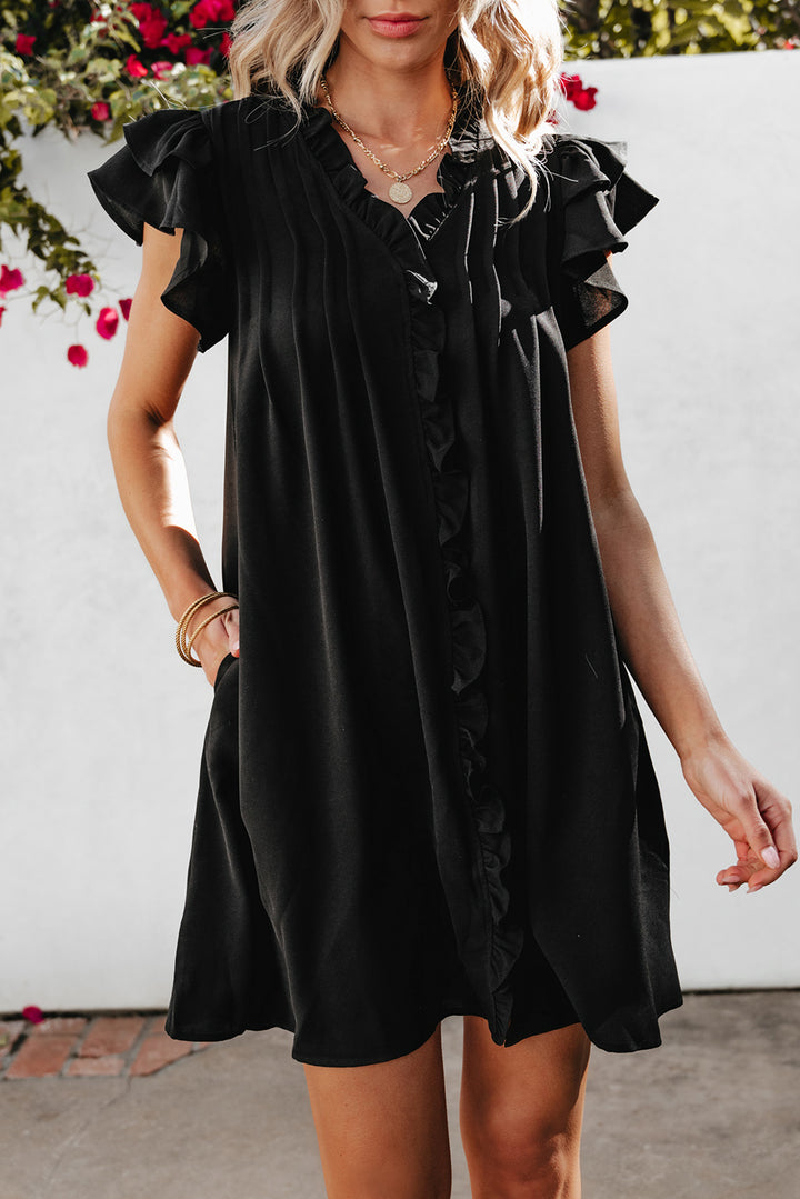 Woman wearing a black dress with ruffled shoulders against a white wall.