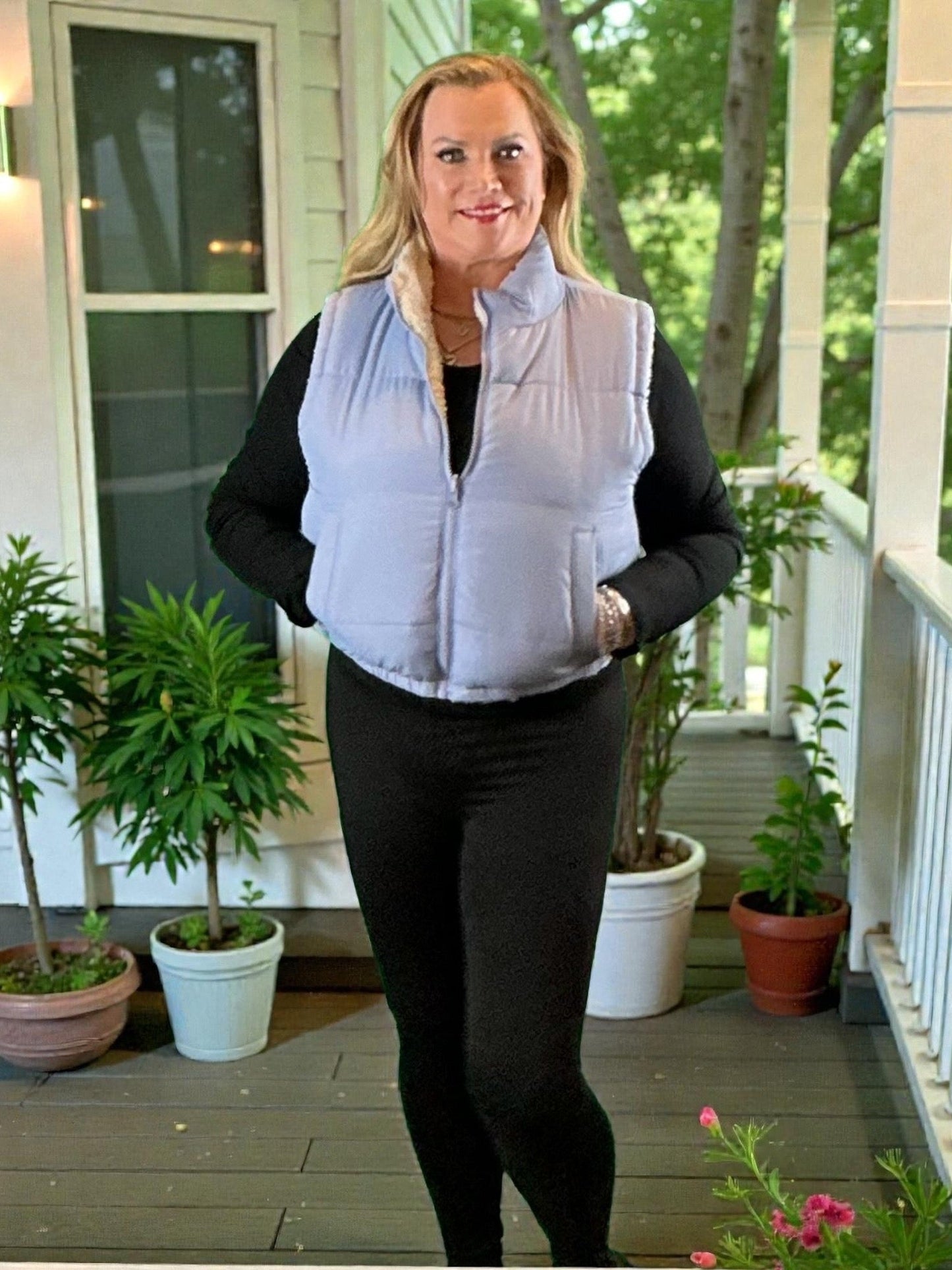 Woman wearing a light gray puffer vest on a porch with plants and a house in the background