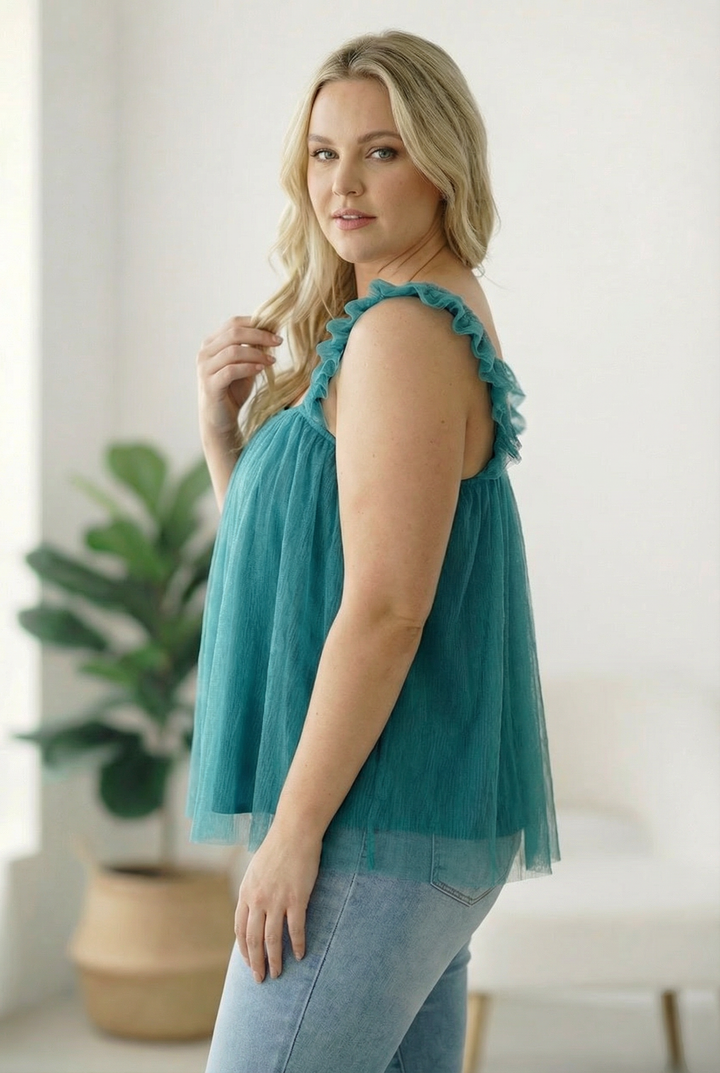 Woman wearing a teal sleeveless top with ruffled shoulders in a bright room.