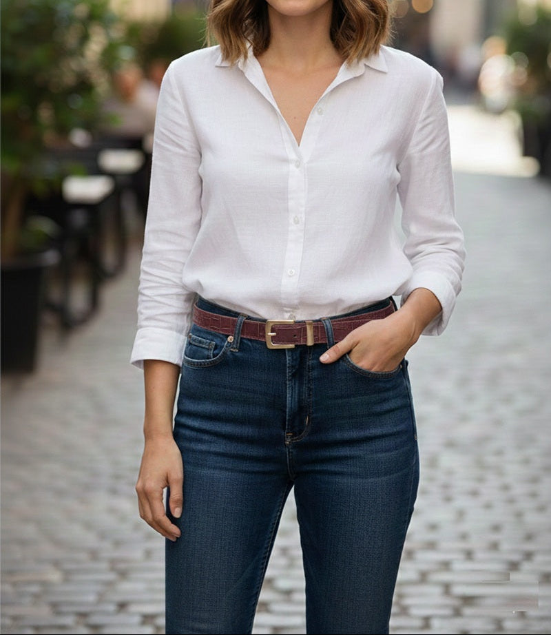 A woman standing on a cobblestone street wearing a white long-sleeve button-up shirt tucked into dark blue jeans with a brown belt.