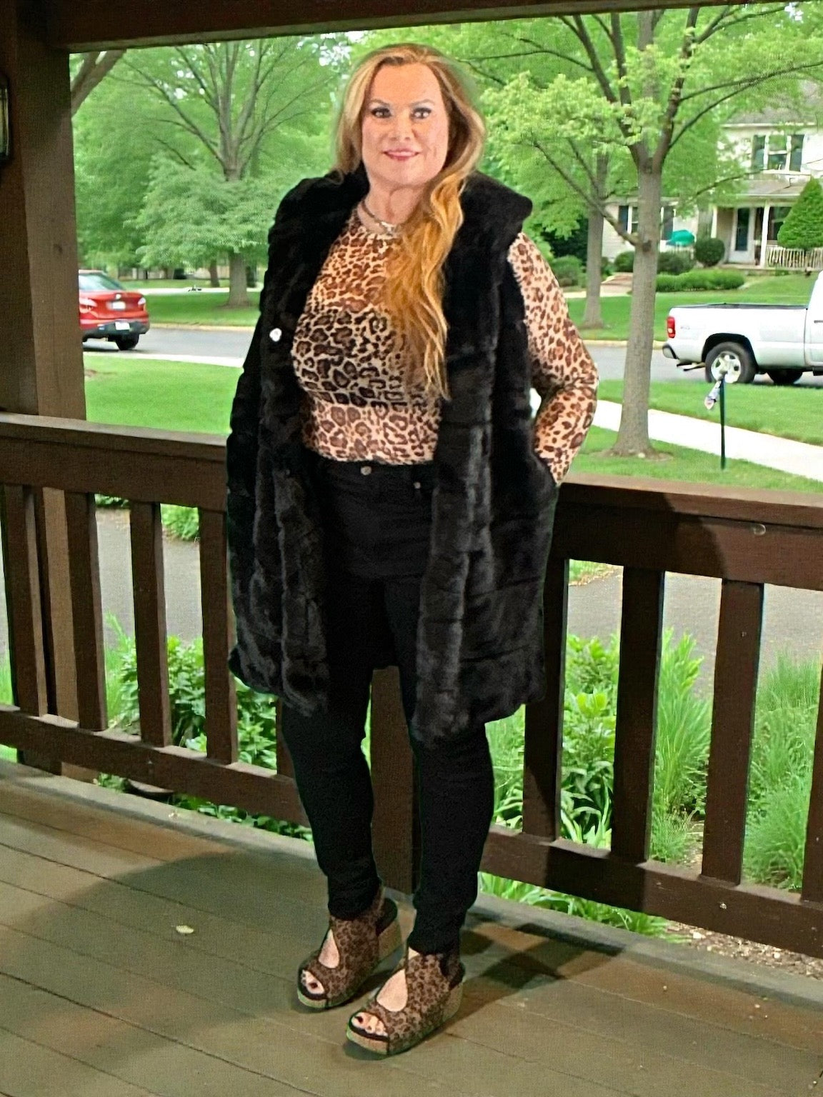 Woman standing on a porch wearing a leopard print top and black fur vest.