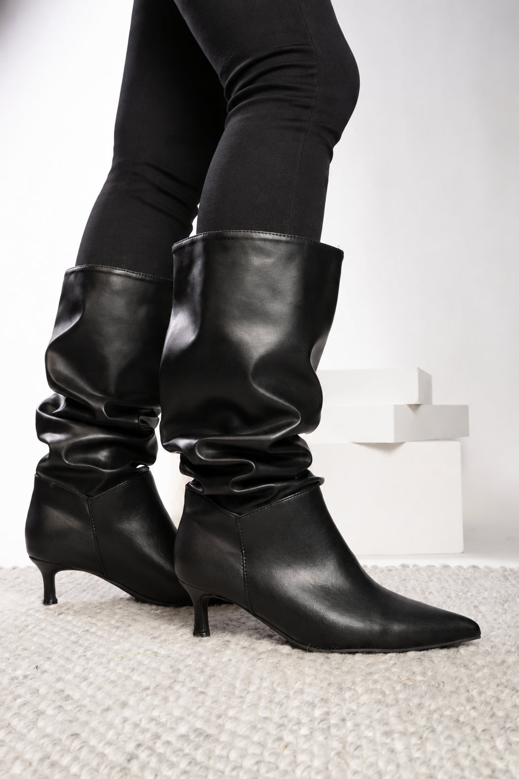 Black slouchy mid-calf boots with kitten heels, worn with light blue jeans on a white textured rug.