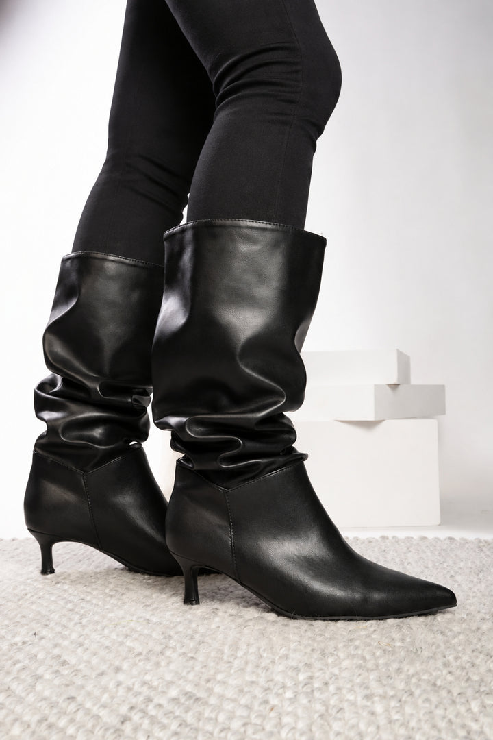 Black slouchy mid-calf boots with kitten heels, worn with light blue jeans on a white textured rug.
