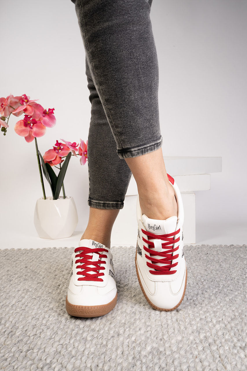 White sneakers with red laces worn by a person, standing on a gray carpet with a white wall and pink flowers in the background.