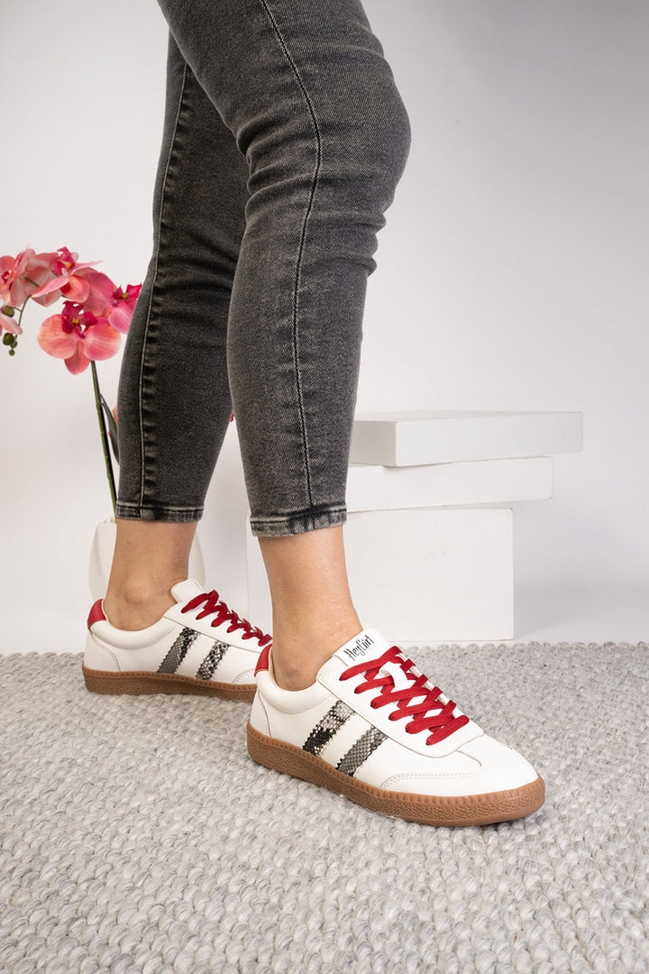 White sneakers with red laces worn with black jeans on a light gray floor.