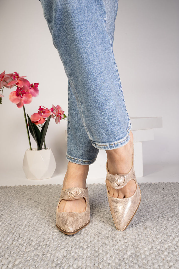 Person wearing gold shoes with bow details and light blue jeans, standing on a carpeted floor with a decorative vase of flowers in the background.