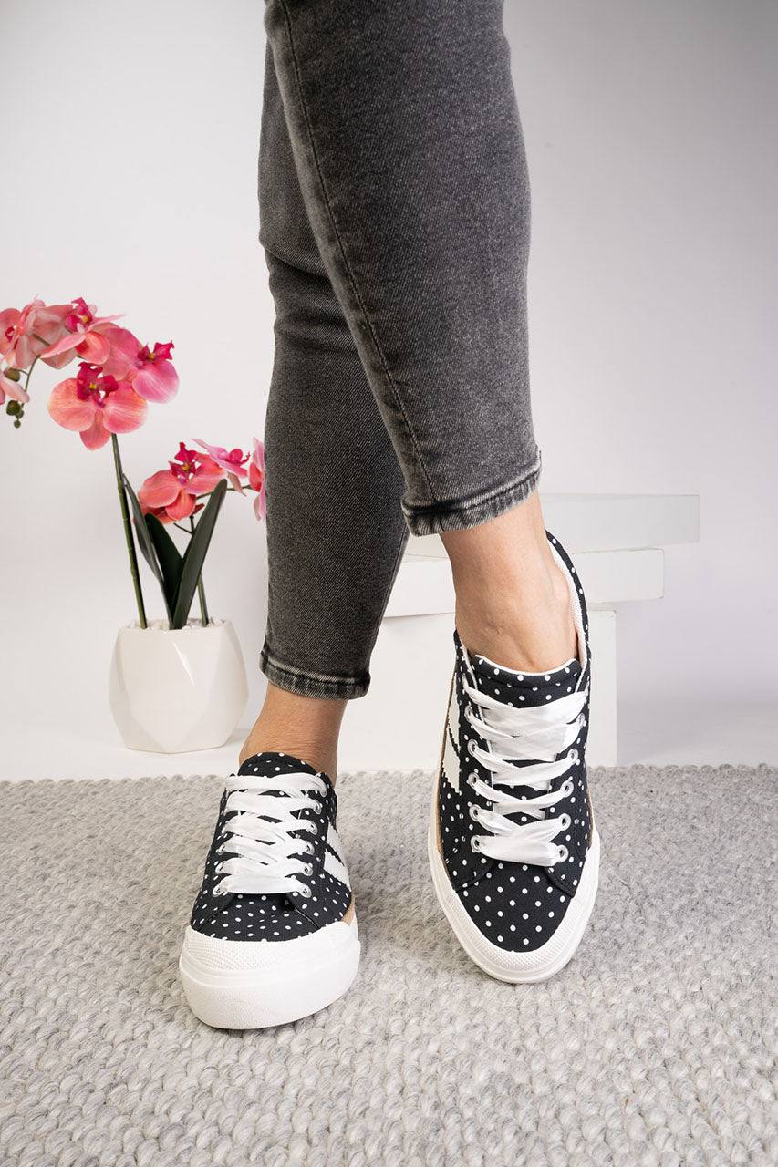 Person wearing black polka dot sneakers with white laces on a neutral background