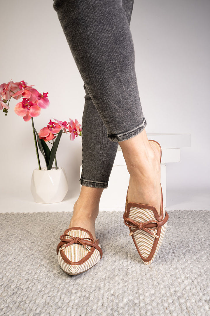 Person wearing brown strappy sandals on a light gray carpet with a white wall and pink flowers in the background.