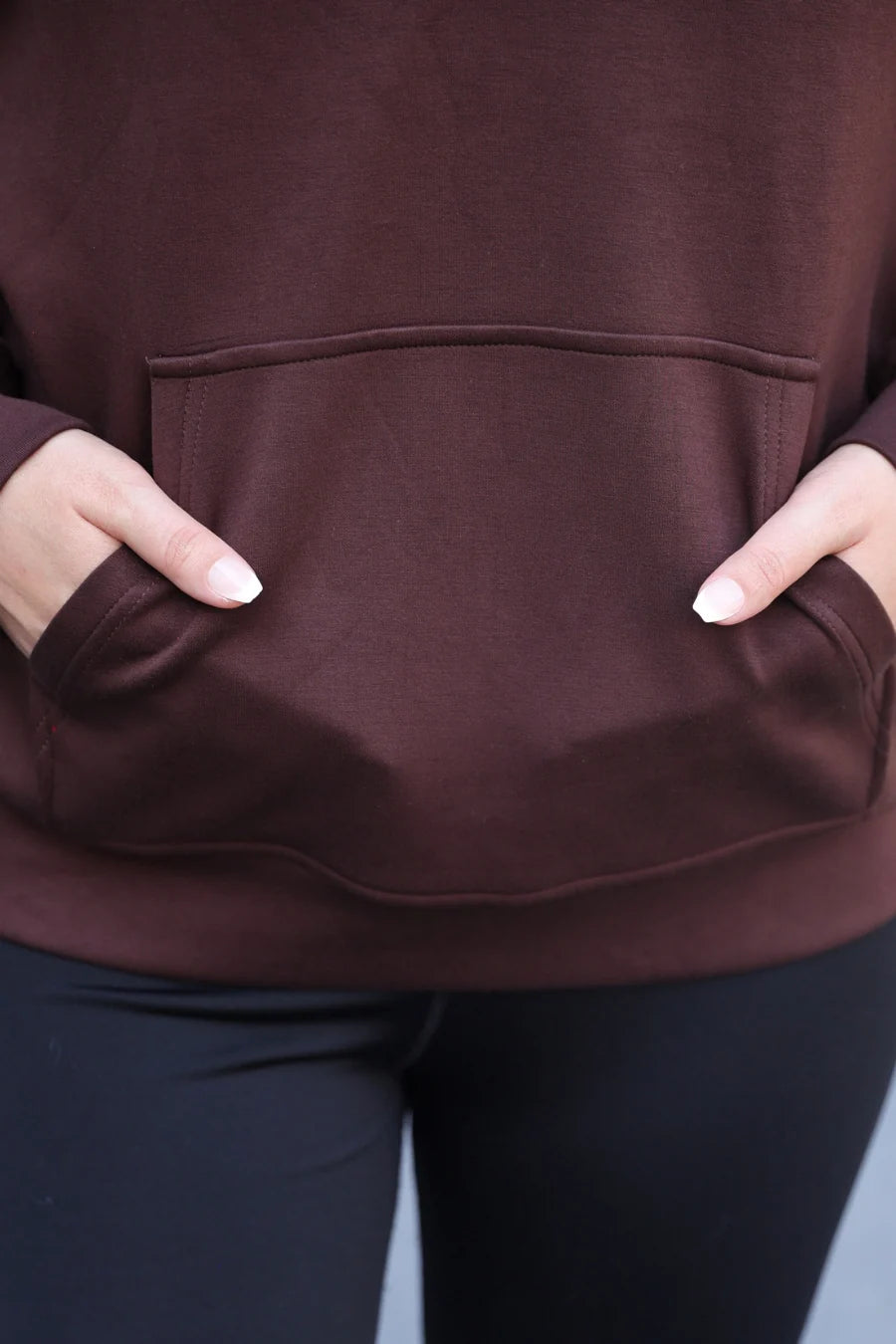 Close-up of a person wearing a chocolate pullover hoodie with a front kangaroo pocket and black leggings, with hands tucked into the pockets.