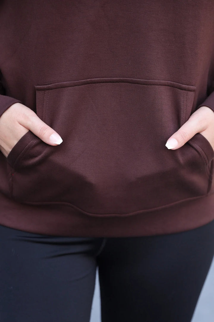 Close-up of a person wearing a chocolate pullover hoodie with a front kangaroo pocket and black leggings, with hands tucked into the pockets.