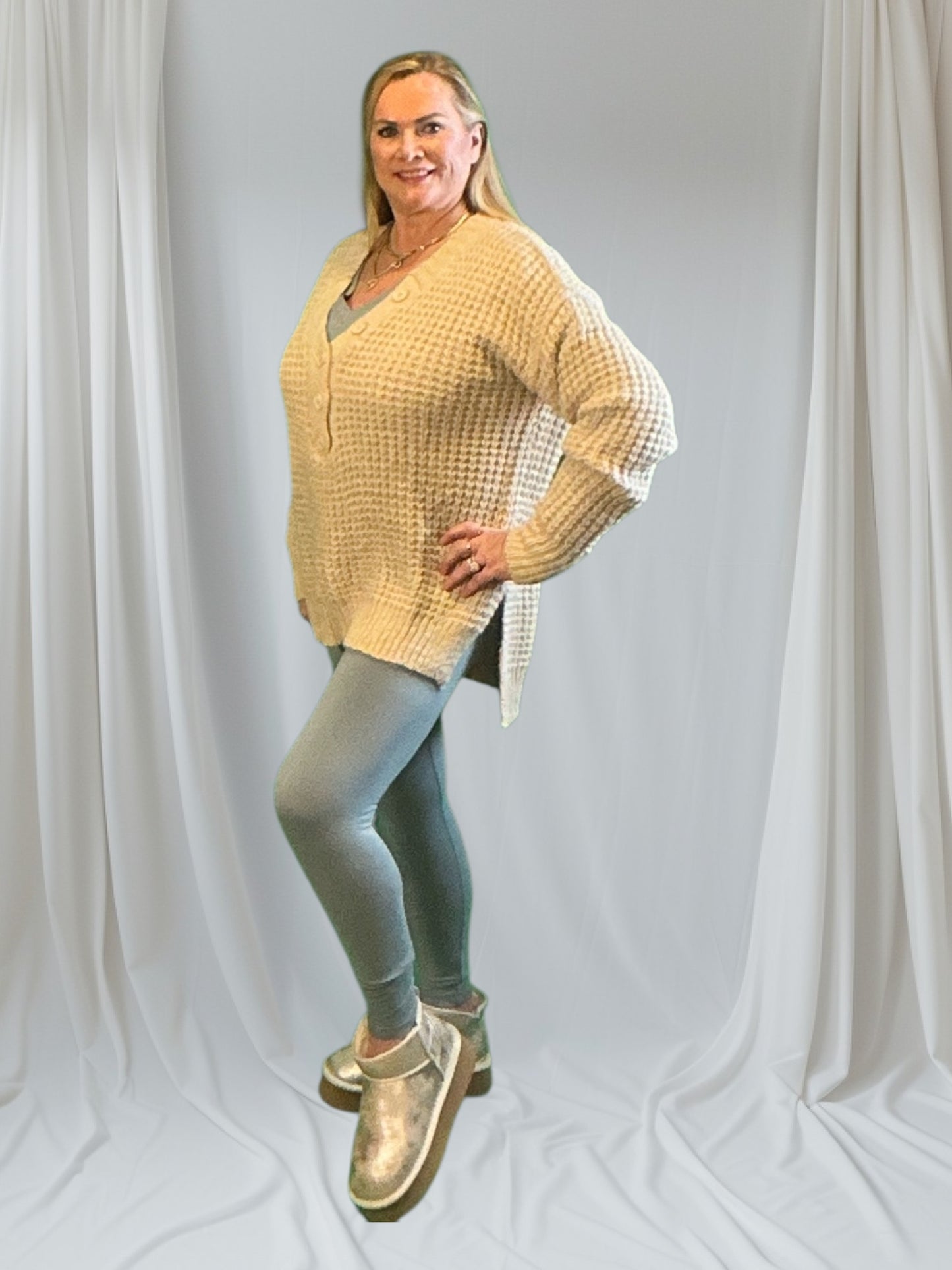Woman wearing a beige cardigan over a checkered shirt, light blue jeans, and gold shoes on a plain background.