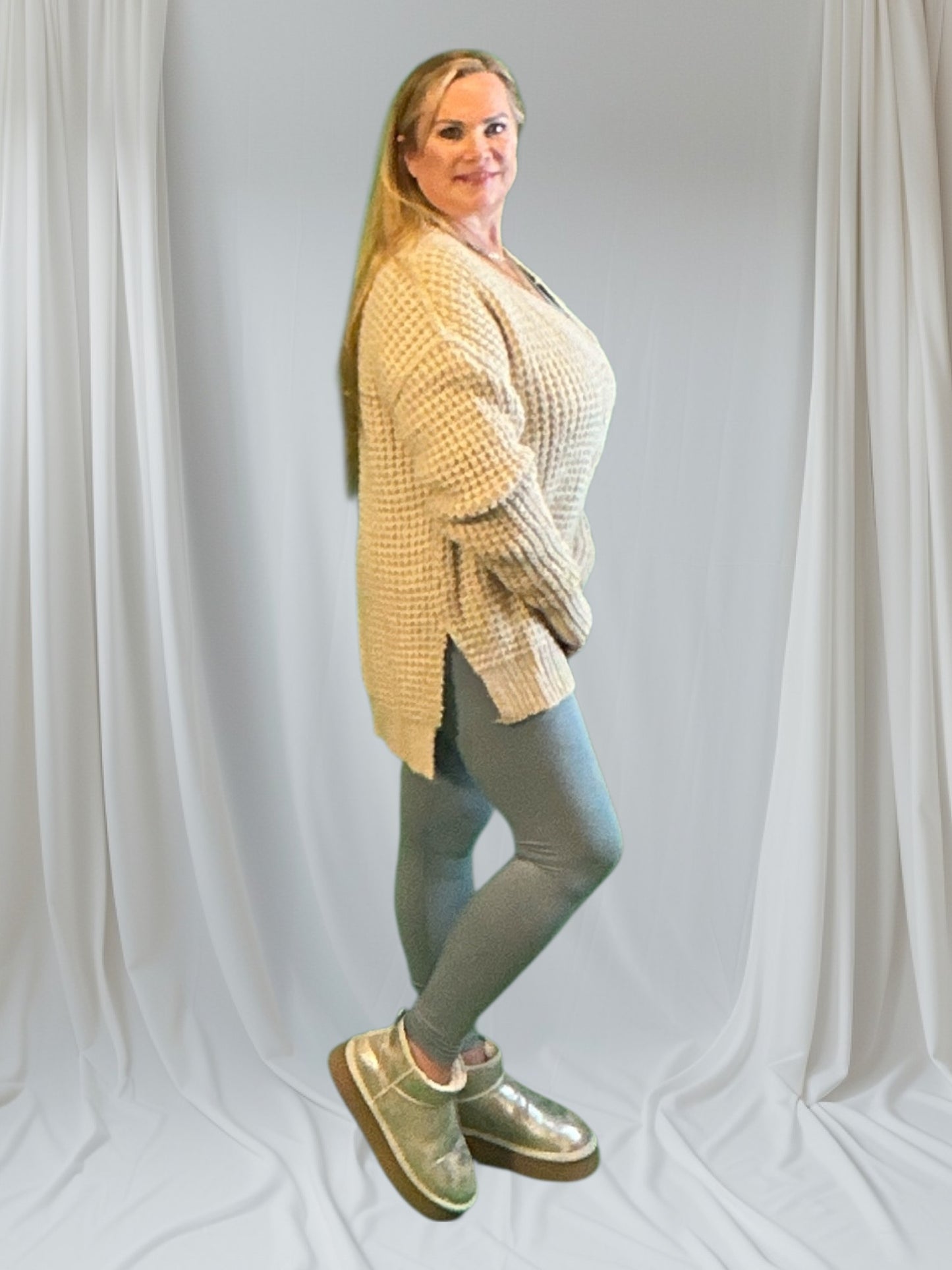 Woman wearing a beige cardigan, gray leggings, and brown boots against a white curtain background