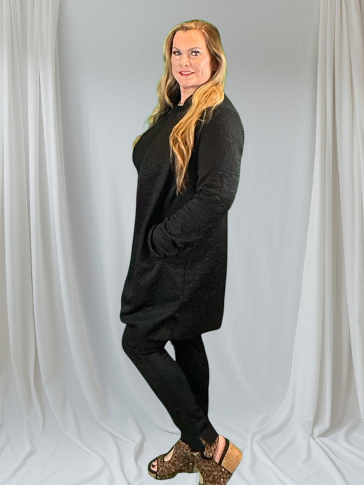 Woman in long blazer with black top and pants against white background.