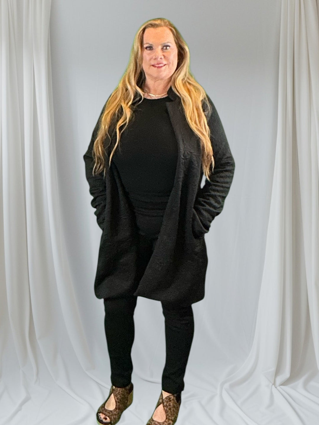 Woman in long blazer with black top and pants against white background.