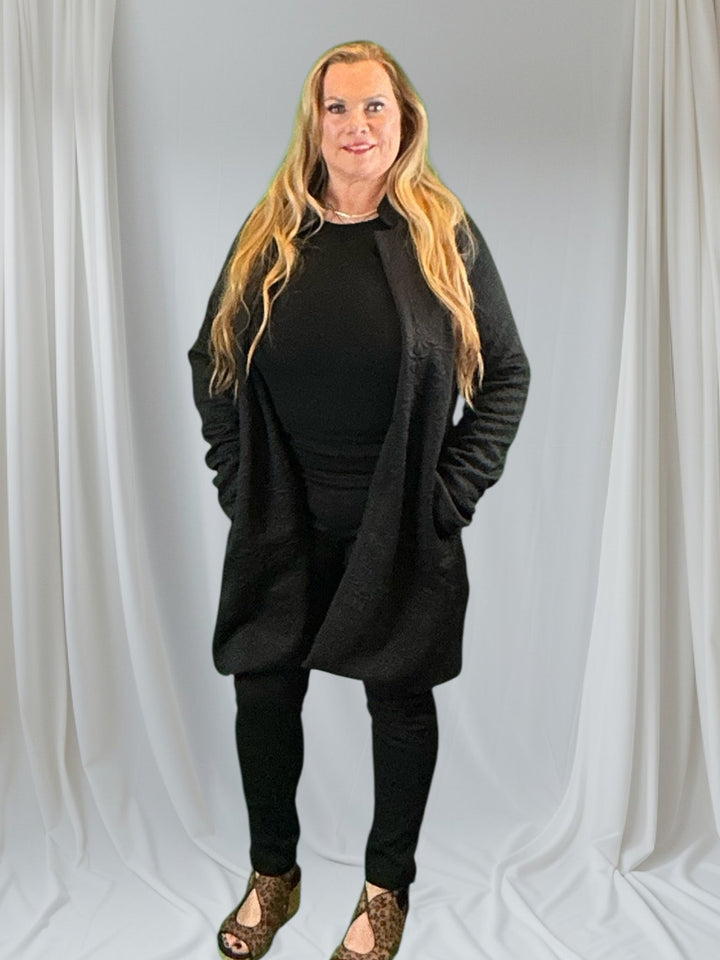 Woman in long blazer with black top and pants against white background.