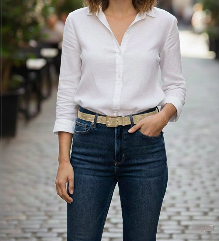 A woman standing on a cobblestone street wearing a white long-sleeve button-down shirt tucked into dark blue jeans with a beige woven belt.