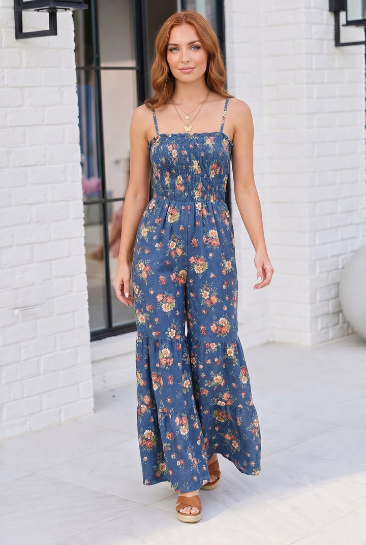 Woman wearing a blue floral jumpsuit standing against a white brick wall.