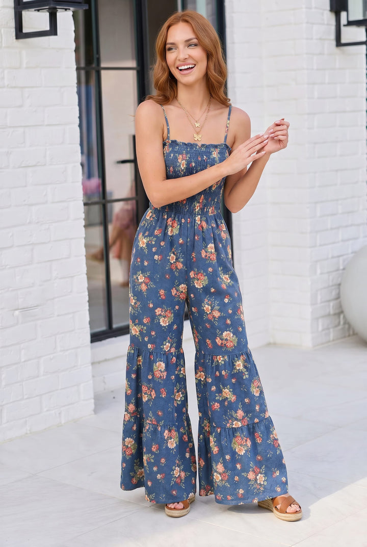 Woman wearing a blue floral jumpsuit standing against a white brick wall.