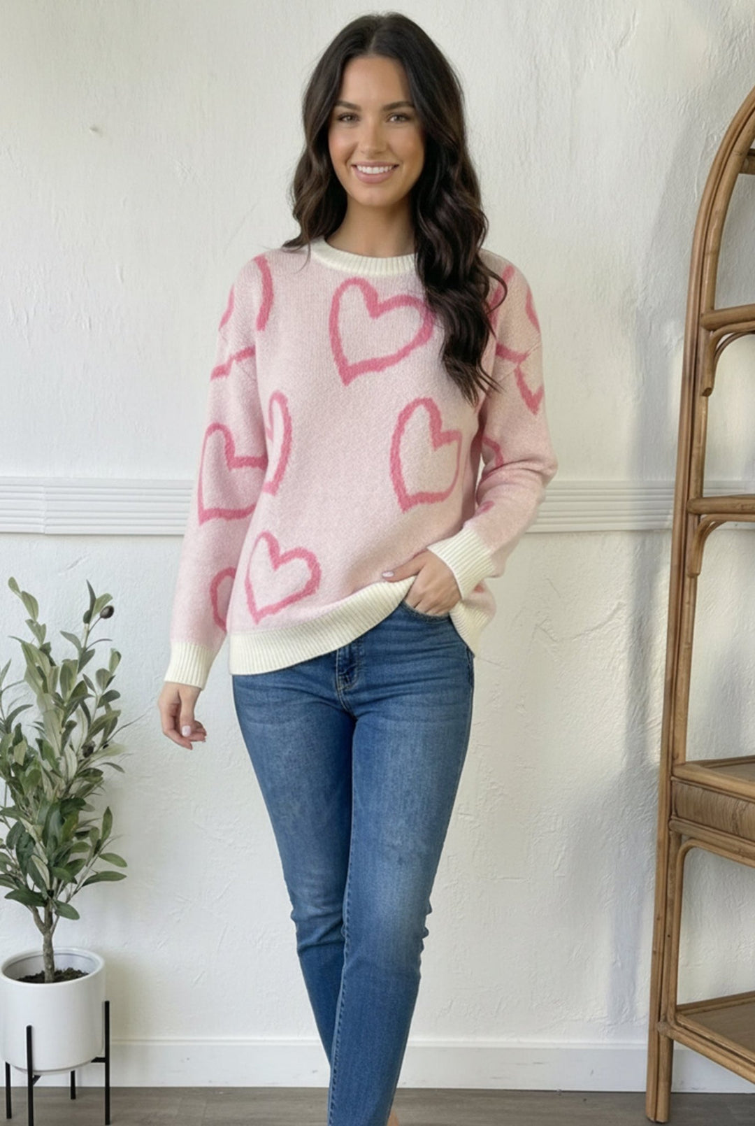 Girl wearing pink sweater with dark pink heart outline and jeans with white background.