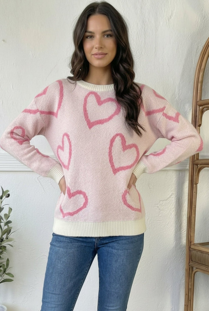 Girl wearing pink sweater with dark pink heart outline and jeans with white background.